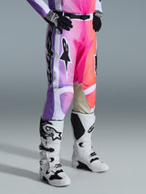 Alpinestars 2026 Racer Air Portl broek, motorcrossbroek, veelkleurige grafische print met paarse, roze en oranje kleurverlopen, gedragen met wit-zwarte motorcrosslaarzen en zwarte handschoenen, voorzien van een race-fit ontwerp en beige lederen panelen aan de binnenkant van de knieën.