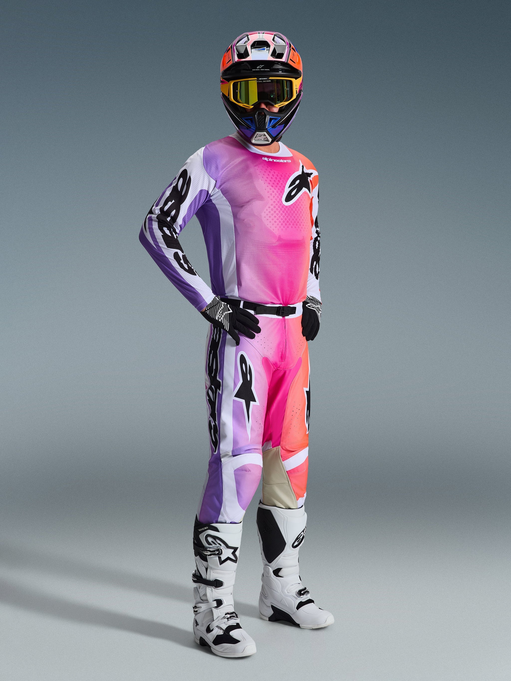 Motorcrossrijder in een volledige race-uitrusting met de Alpinestars 2026 Racer Air Portl-broek, een opvallend meerkleurig ontwerp in roze, paars en oranje met witte accenten, bijpassende jersey met lange mouwen, zwarte helm met gele bril en witte laarzen, een gecoördineerde outfit voor high-performance off-road racen.