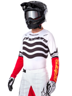 Alpinestars Limited Edition Techstar Deep Jersey, motorcrosser met een wit shirt met zwarte golvende strepen en rode onderkant van de mouwen, gecombineerd met een zwarte helm, spiegelende crossbril, wit-zwarte handschoenen en witte crossbroek, performance motorcross outfit.