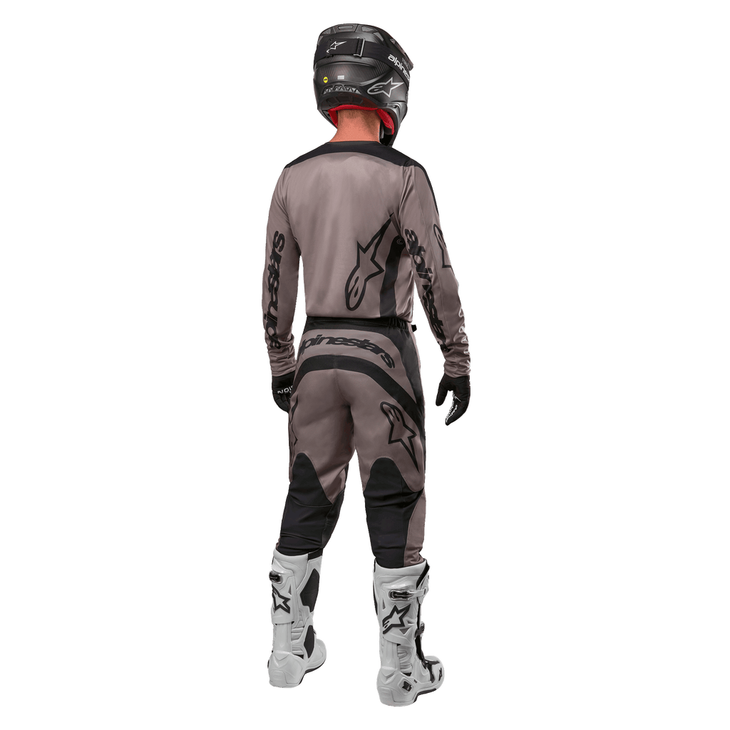 Alpinestars 2024 Fluid Lurv Jersey, motocross jersey, Mud Black, achteraanzicht van een motorcrossrijder in bijpassende race-uitrusting, zwarte helm en witte laarzen, met opvallende zwarte Alpinestars-logo's en een sportieve pasvorm voor off-road prestaties.