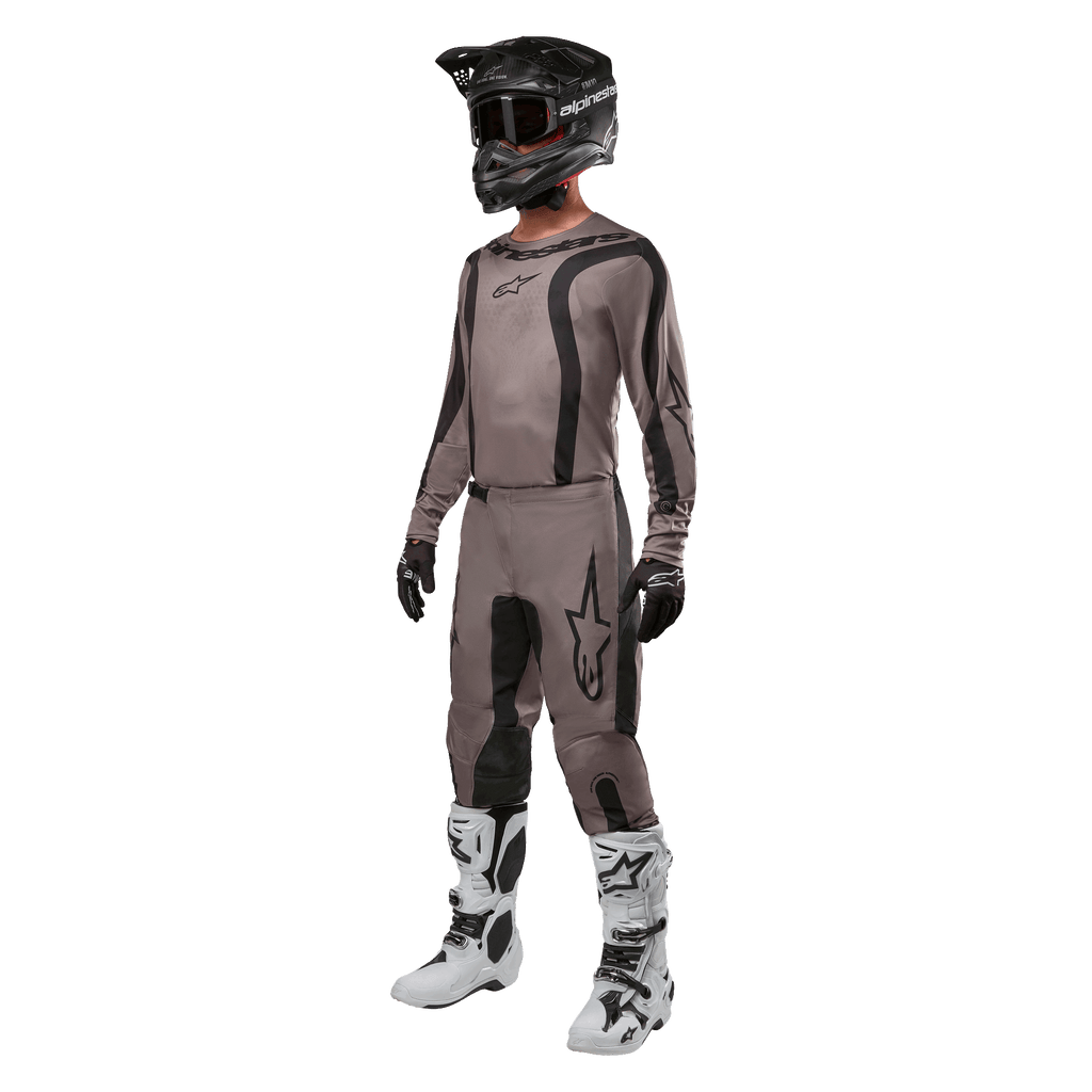 Motorcrossrijder in volledige race-uitrusting met de Alpinestars 2024 Fluid Lurv Jersey in Mud Black, gecombineerd met bijpassende broek, zwarte helm met goggle, wit-zwarte laarzen en zwarte handschoenen, gecoördineerde outfit voor off-road racen