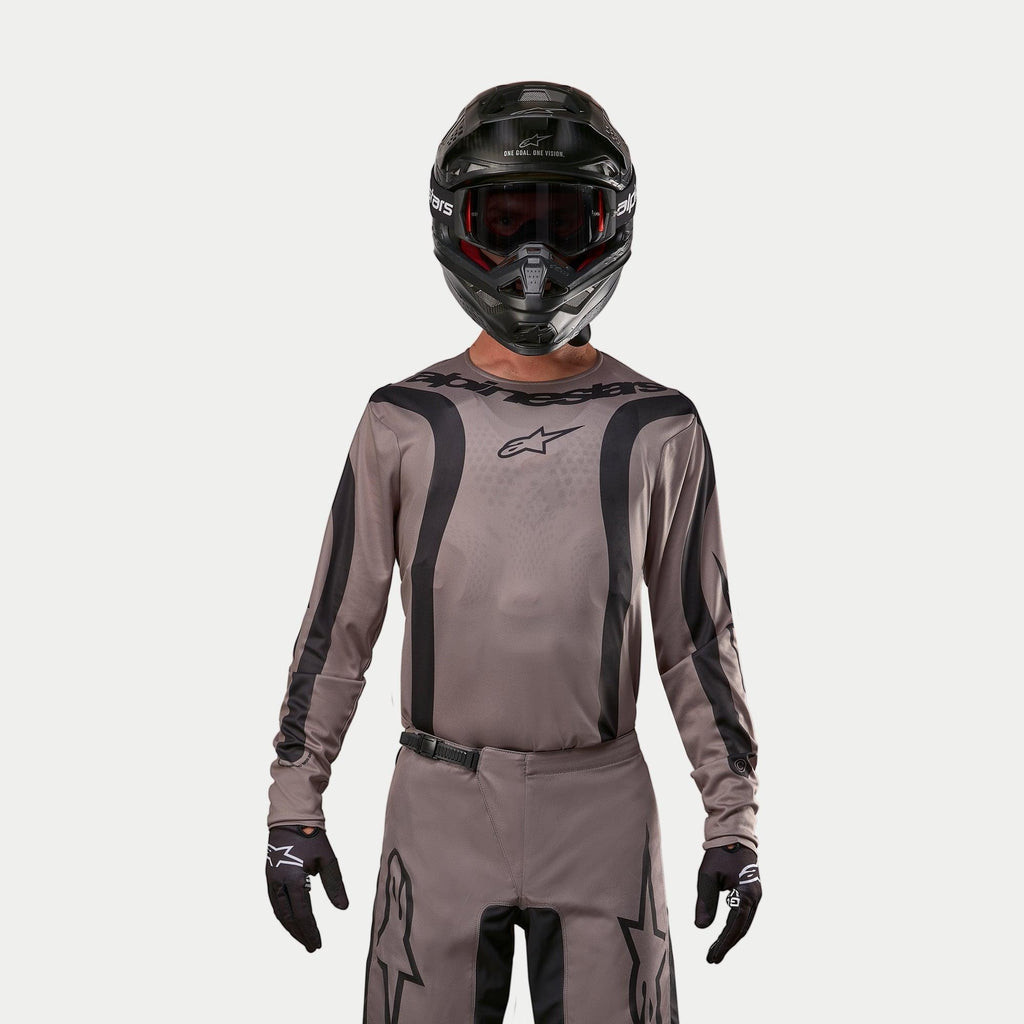 Motorcrosser in de Alpinestars 2024 Fluid Lurv Jersey in Mud Black, een aardse grijsbruine stof met zwarte grafische strepen en een sterlogo op de borst, gecombineerd met een zwarte helm, rode crossbril en zwarte handschoenen voor een gecoördineerde off-road race-look