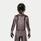 Motorcrosser in de Alpinestars 2024 Fluid Lurv Jersey in Mud Black, een aardse grijsbruine stof met zwarte grafische strepen en een sterlogo op de borst, gecombineerd met een zwarte helm, rode crossbril en zwarte handschoenen voor een gecoördineerde off-road race-look