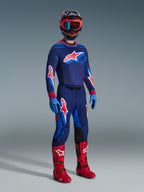 Motocross rider wearing complete racing gear, 2026 Supertech Vista Jersey in Dark Navy with Hot Coral and Cobalt accents, matching motocross pants, multi-colored helmet with red goggles, bright red boots, blue gloves, coordinated outfit for off-road racing