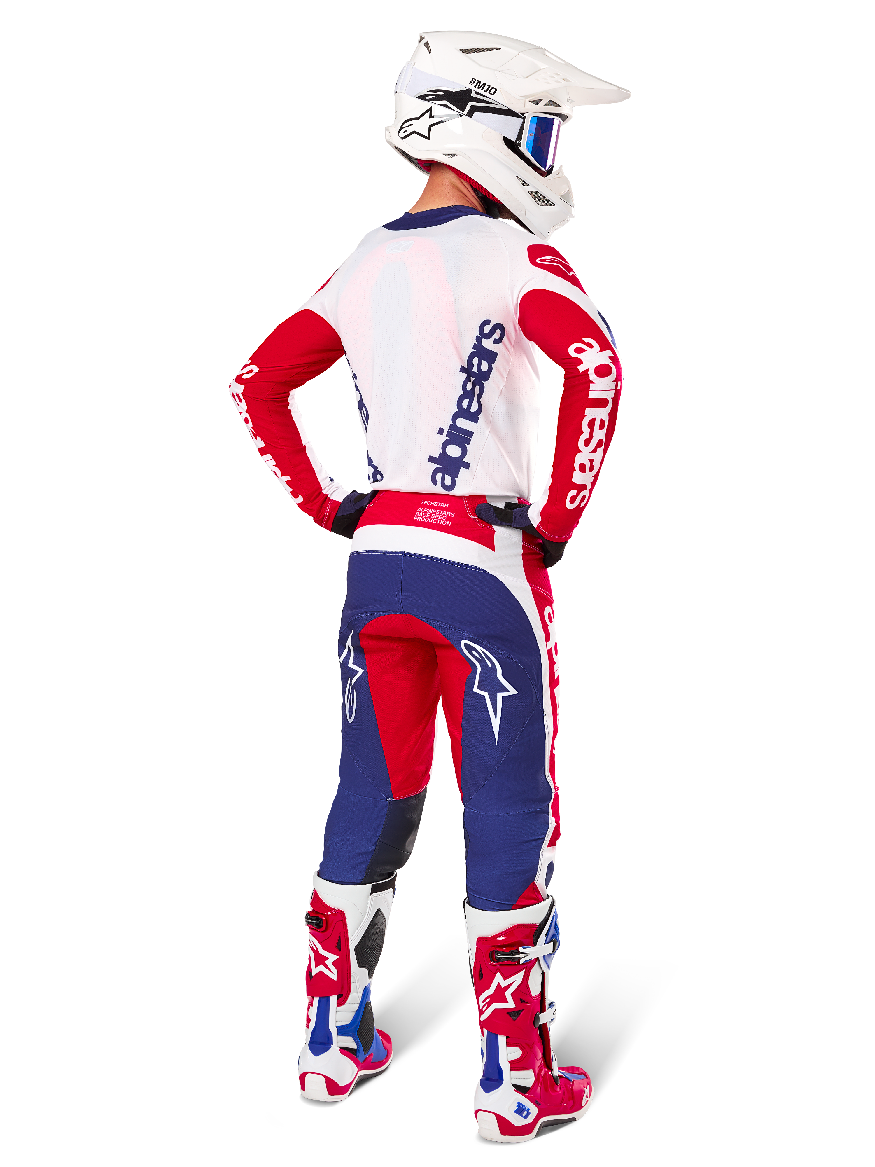 Motorcrosser van achteren gezien in de Alpinestars Limited Edition Techstar Fortitude Jersey en uitrustingsset, Bright Red White Blue kleurstelling, met witte Alpinestars-branding op de rode mouwen en een navy logo op de witte achterkant, gecombineerd met een witte motorcrosshelm en bijpassende racelaarzen.
