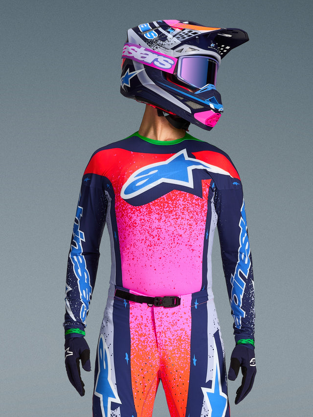 Motocross rider wearing the Alpinestars Limited Edition Supertech Records Jersey, vibrant pink and navy design with orange speckle patterns and a light blue star logo, matching helmet and goggles, professional racing gear.