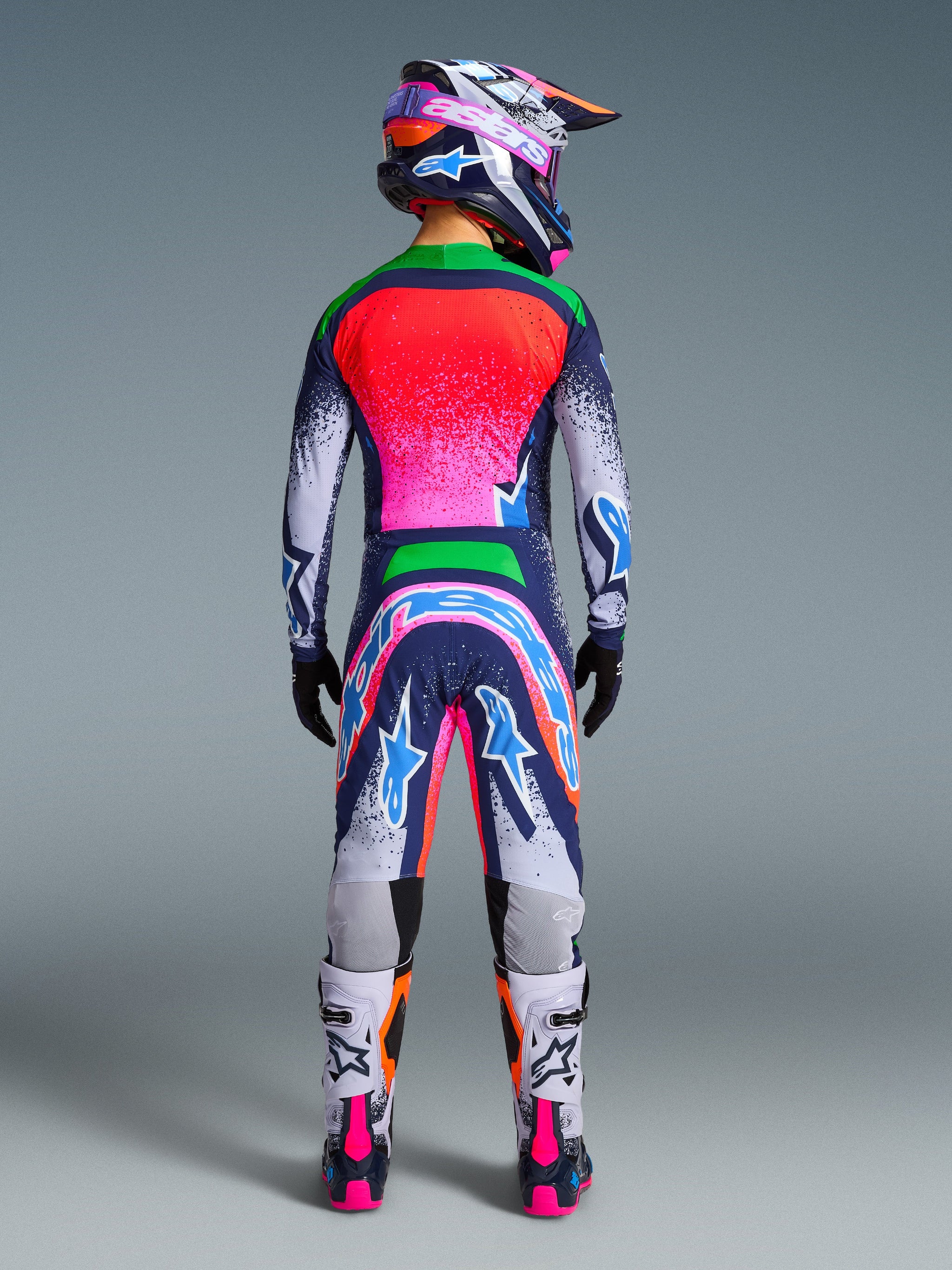 Achteraanzicht van een motorcrosser in de volledige Alpinestars Limited Edition Supertech Records-collectie, inclusief een meerkleurige jersey en broek met opvallende marineblauwe, neonroze en oranje accenten, bijpassende helm, zwarte handschoenen en witte motorcrosslaarzen, gecoördineerde uitrusting voor professionele races.