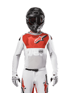 Alpinestars 2024 Supertech Ward Jersey, motorcross shirt in White Hot Orange, wit stretchgaas jersey met een opvallende rode borstgrafiek en logo, zwarte helm met donkere bril, witte handschoenen, professionele uitrusting voor off-road prestaties