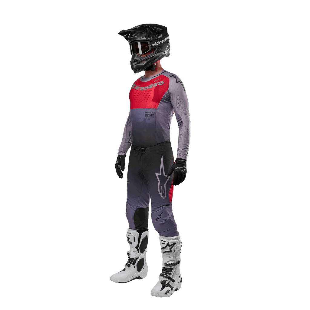 Motorcrossrijder in volledige race-uitrusting, Alpinestars 2024 Supertech Dade Jersey in Iron Red Berry en grijs, bijpassende grijze broek met rode accenten, zwarte helm met crossbril, witte en zwarte laarzen, zwarte handschoenen, gecoördineerde outfit voor off-road racen