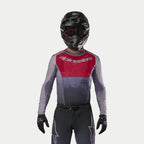 Alpinestars 2024 Supertech Dade Jersey in Iron Red Berry, motocross jersey met een rood borstpaneel en grijze mouwen met zilveren logo's, gedragen door een motocrosser met een bijpassende broek, zwarte helm en zwarte handschoenen, professionele off-road race-uitrusting
