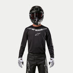 Alpinestars 2024 Fluid Graphite Jersey, zwart en zilveren motorcross jersey, gedragen door een rijder met een zwarte helm en rode bril, voorzien van zilveren Alpinestars branding op de borst, gecombineerd met een zwarte broek en witte handschoenen voor off-road prestaties