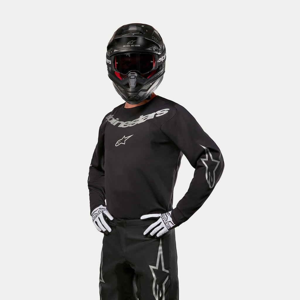 Motorcrosser die de Alpinestars 2024 Fluid Graphite Jersey draagt, zwart en zilver, gecombineerd met een zwarte crossbroek, zwarte helm met rood getinte bril en wit-zwarte handschoenen, voorzien van zilveren merklogo's op de borst en mouwen.