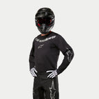 Motorcrosser die de Alpinestars 2024 Fluid Graphite Jersey draagt, zwart en zilver, gecombineerd met een zwarte crossbroek, zwarte helm met rood getinte bril en wit-zwarte handschoenen, voorzien van zilveren merklogo's op de borst en mouwen.