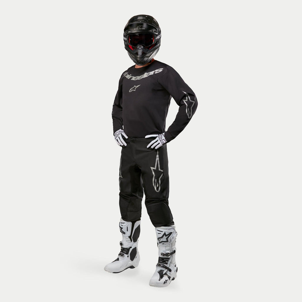 Motocrossrijder in Alpinestars 2024 Fluid Graphite Jersey in zwart en zilver, bijpassende zwarte broek met zilveren ster-logo's, wit-zwarte laarzen, wit-zwarte handschoenen en een zwarte helm met rode crossbril, volledige off-road race-uitrusting