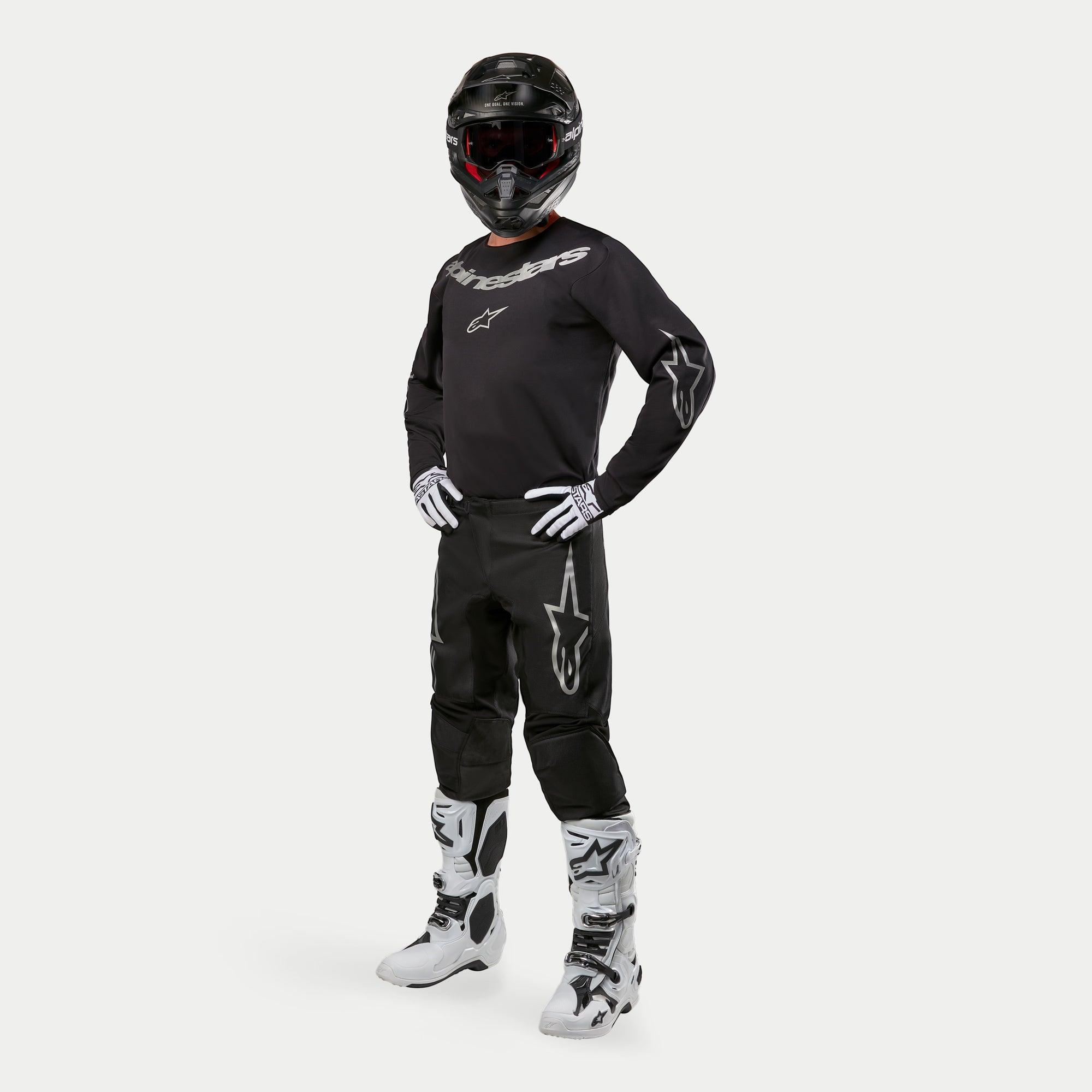 Motocrossrijder in Alpinestars 2024 Fluid Graphite Jersey in zwart en zilver, bijpassende zwarte broek met zilveren ster-logo's, wit-zwarte laarzen, wit-zwarte handschoenen en een zwarte helm met rode crossbril, volledige off-road race-uitrusting