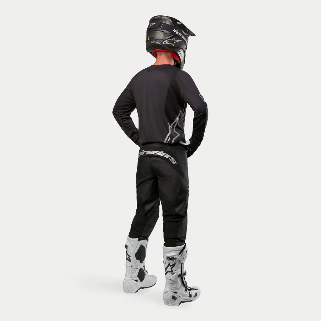 Alpinestars 2024 Fluid Graphite Jersey in zwart-zilver, motocrossrijder achteraanzicht, atletische pasvorm jersey met lange mouwen, gecombineerd met een zwarte crossbroek, zwarte helm en wit-zwarte laarzen, complete professionele uitrusting voor off-road racing.