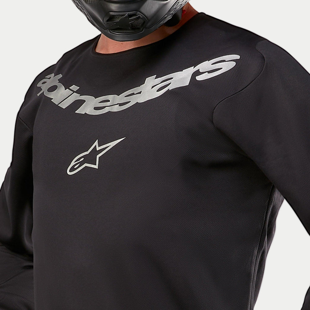 Alpinestars 2024 Fluid Graphite Jersey, motocross jersey, zwart en zilver, close-up detail van de borst met zilveren Alpinestars-branding en sterlogo, ademende poly-stof constructie, gedragen door een rijder met een zwarte helm