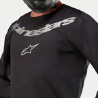 Alpinestars 2024 Fluid Graphite Jersey, motocross jersey, zwart en zilver, close-up detail van de borst met zilveren Alpinestars-branding en sterlogo, ademende poly-stof constructie, gedragen door een rijder met een zwarte helm