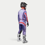 Alpinestars 2024 Techstar Ocuri motorcross outfit, compleet racetenue met paarse jersey en broek, fel oranje logo's, witte laarzen en zwarte helm, achteraanzicht van een rijder in volledige uitrusting