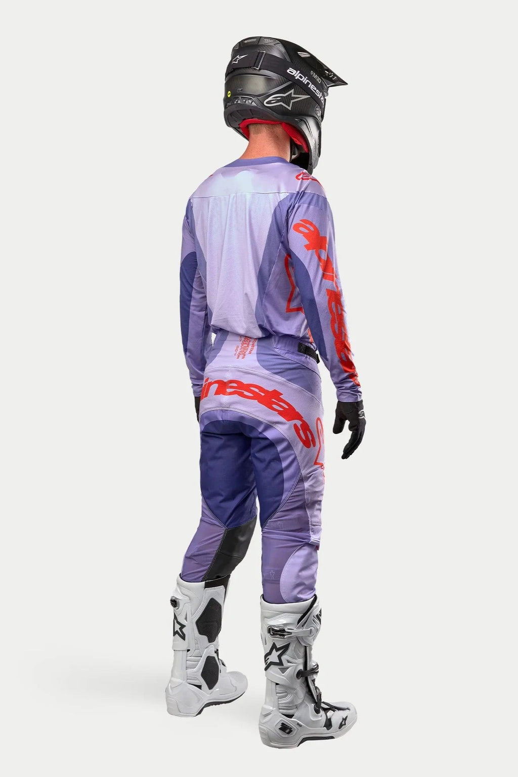 Motorcrosser in complete race-uitrusting van achteren gezien, paars Alpinestars 2024 Techstar Ocuri Jersey met feloranje branding, bijpassende paarse broek met oranje details, zwarte helm, witte en zwarte laarzen, zwarte handschoenen, professionele off-road racing kit.
