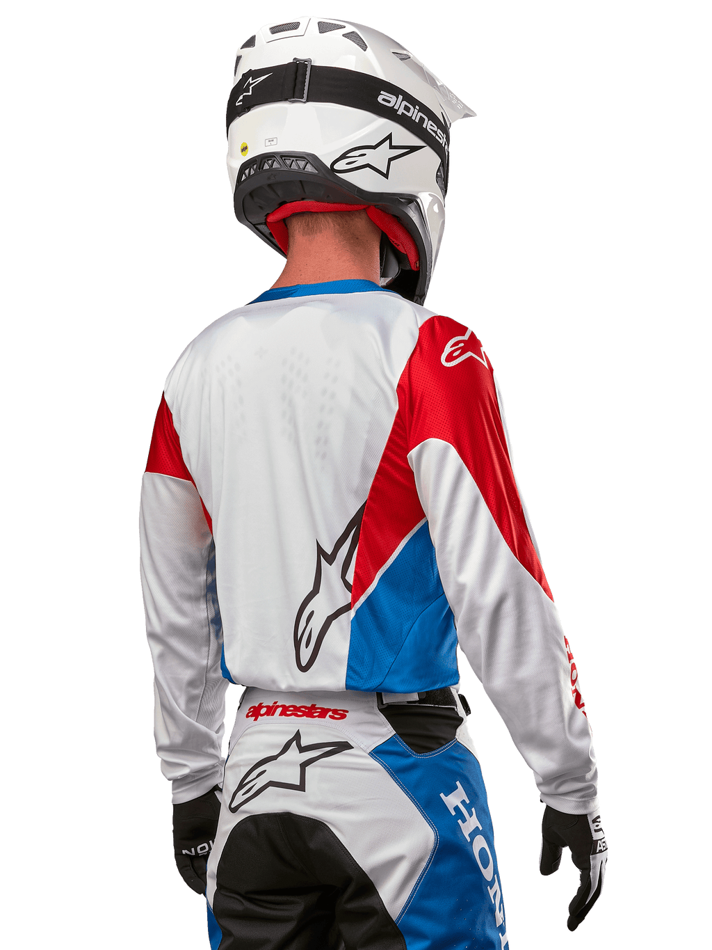Motorcrosser in het Alpinestars Honda Racer Iconic Jersey, achteraanzicht, wit met felblauwe en felrode panelen, opvallend Alpinestars-logo op de rug, gecombineerd met een witte motorcrosshelm en bijpassende crossbroek