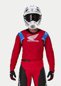 Motocross-rijder die de Alpinestars Honda Racer Iconic Jersey draagt, felrood met blauwe en witte accenten en Honda-branding, gecombineerd met een witte en blauwe helm, donkere bril en bijpassende rode en zwarte motorcrossbroek, prestatiegerichte uitrusting voor off-road rijden