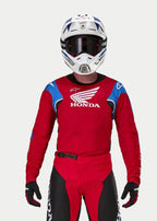 Motocross-rijder die de Alpinestars Honda Racer Iconic Jersey draagt, felrood met blauwe en witte accenten en Honda-branding, gecombineerd met een witte en blauwe helm, donkere bril en bijpassende rode en zwarte motorcrossbroek, prestatiegerichte uitrusting voor off-road rijden