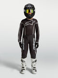 Alpinestars jeugd Fluid Apex V2 jersey en gear set, zwart-zilveren motocross-outfit met witte motorlaarzen en helm.