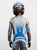Alpinestars Jeugd Fluid Drag Jersey 2027, motorcross-jersey voor kinderen, grijs en blauw, ademende polyfabric constructie, achteraanzicht van een jonge rijder met helm, professionele off-road race-uitrusting
