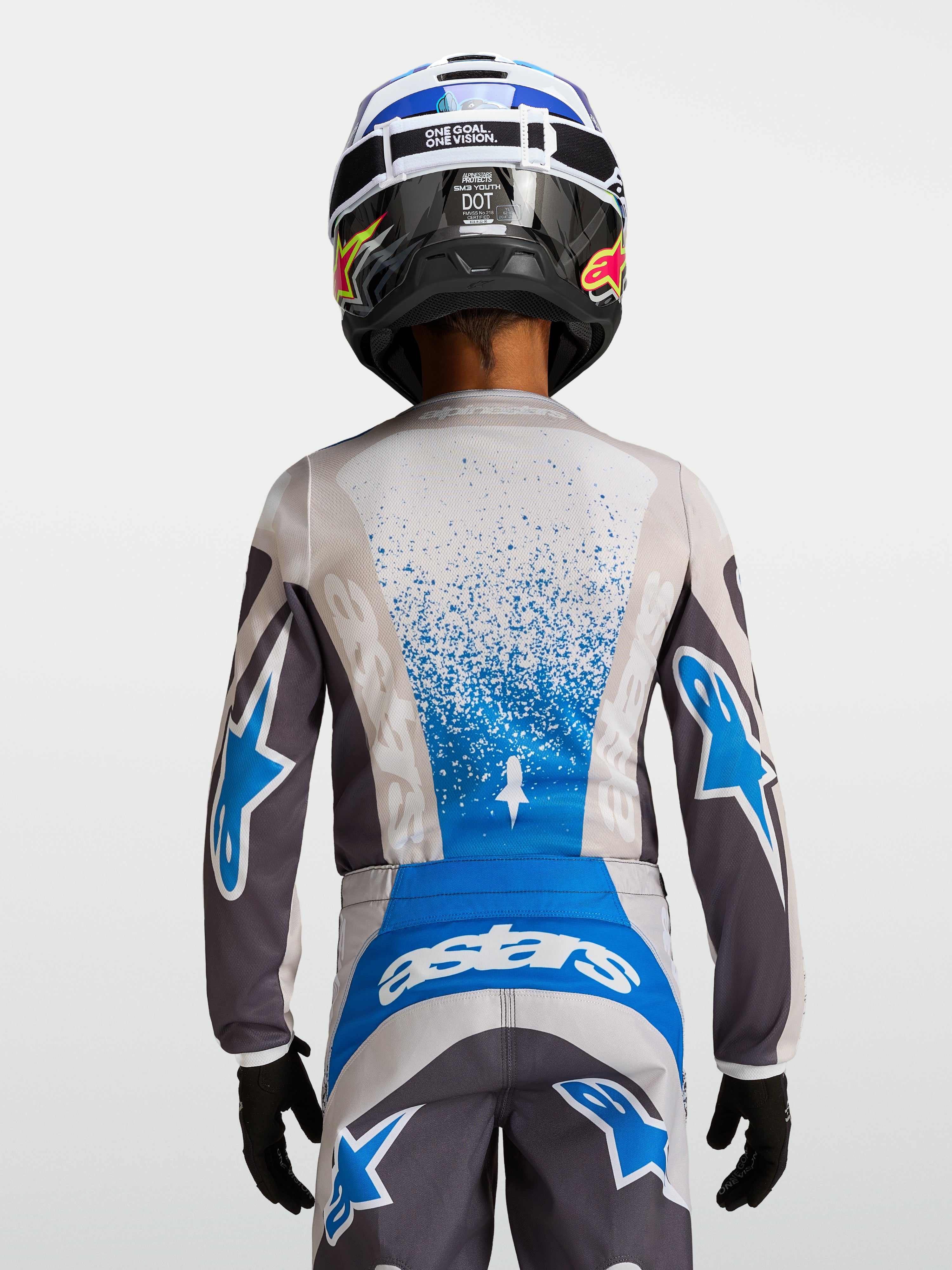 Alpinestars Jeugd Fluid Drag Jersey 2027, motorcross-jersey voor kinderen, grijs en blauw, ademende polyfabric constructie, achteraanzicht van een jonge rijder met helm, professionele off-road race-uitrusting