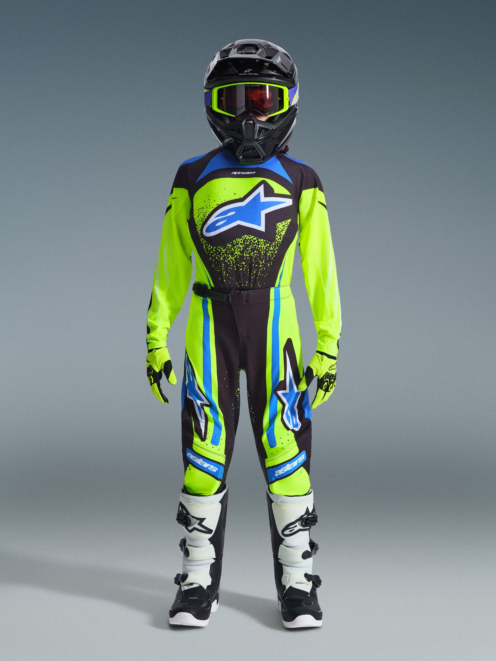 Jeugd motocrossrijder in volledige race-uitrusting, 2026 Jeugd Supertech Nomur Jersey in neongeel en donkerblauw met een blauw Alpinestars-logo, bijpassende broek, wit-zwarte laarzen, neongele handschoenen en een zwarte helm met crossbril, professionele off-road race-outfit.