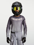 Alpinestars Jeugd Fluid Amber jersey, ademend motorcrossjersey in grijs en zand, gedragen door een jonge rijder met helm.