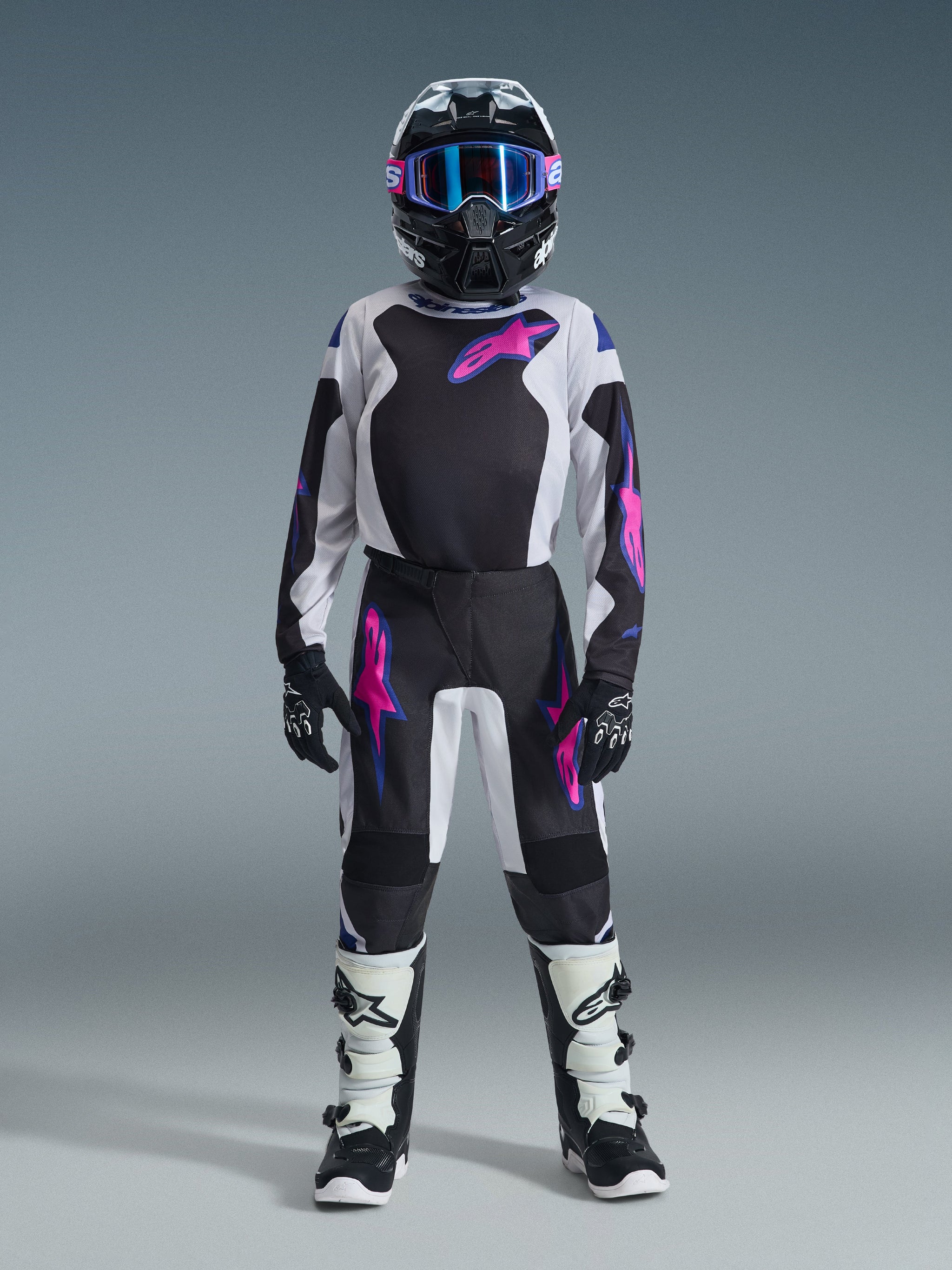 Young motocross rider wearing the Alpinestars 2026 Youth Fluid Grid Jersey in Black, Light Gray, and Purple, paired with matching motocross pants, black helmet with pink accents and blue goggles, black and white boots, and black gloves, complete professional off-road racing gear set