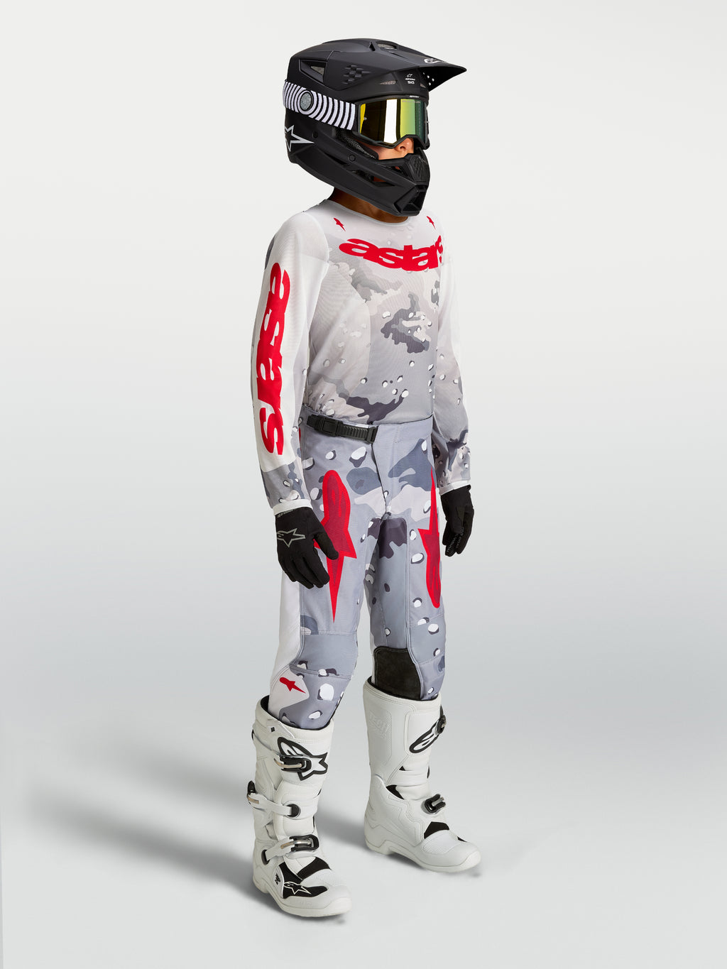 Alpinestars jeugd Fluid Lone gearset, grijze camo en rode motorcrossuitrusting op een rijder met witte motorlaarzen.
