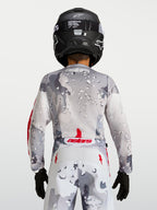 Alpinestars Jeugd Fluid Lone Jersey 2027, grijze camouflage motorcrossjersey, achteraanzicht met rode accenten en helm.