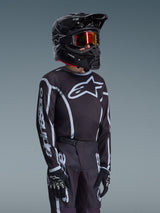 Alpinestars 2026 Youth Fluid Apex Jersey in zwart en grijs, gedragen door een jonge rijder met een bijpassende zwarte helm en getinte bril, met opvallende Alpinestars-branding op de borst en mouwen, onderdeel van een gecoördineerde motorcross-uitrusting.