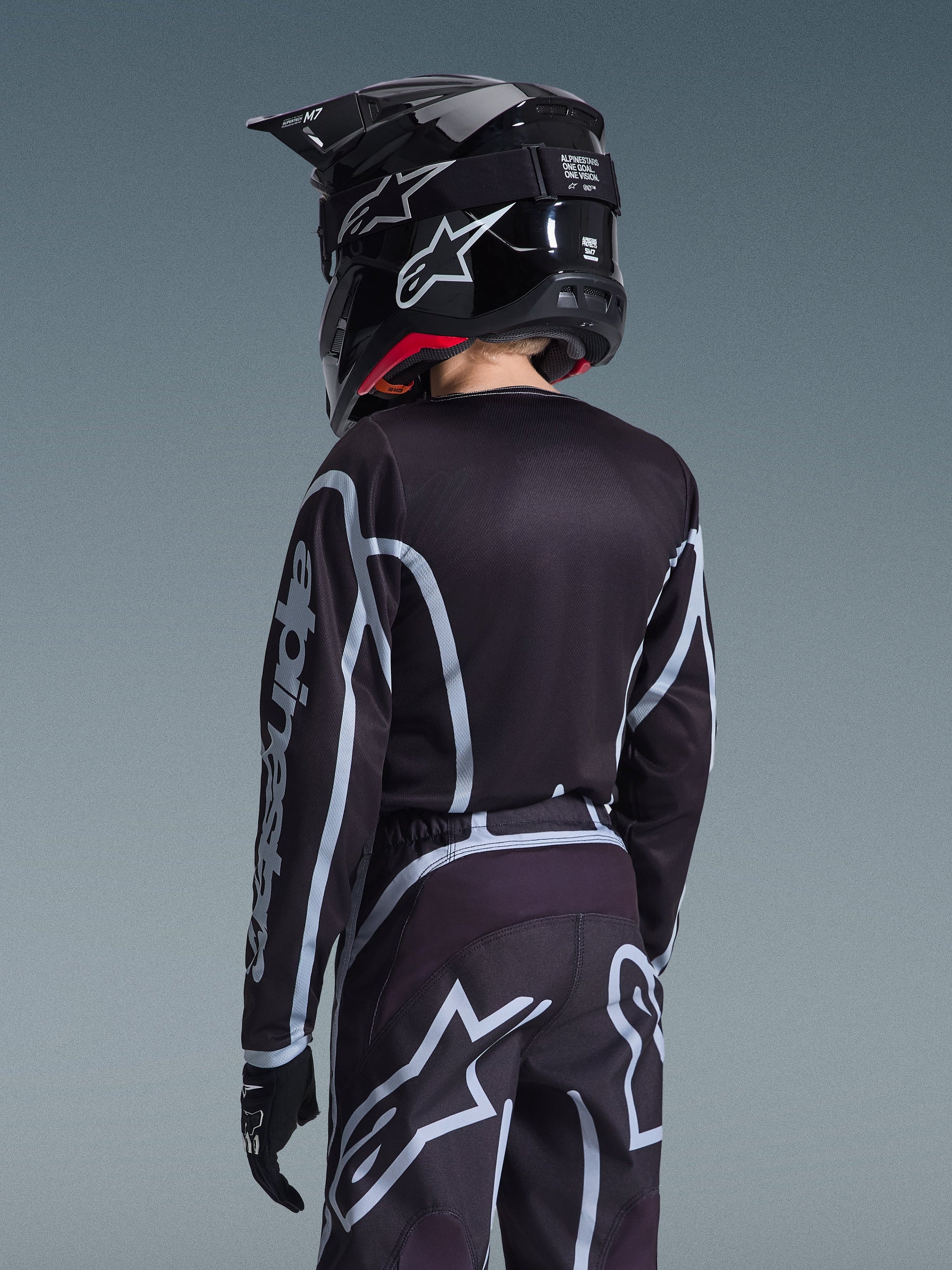 Achteraanzicht van een jonge motocrosser die de Alpinestars 2026 Youth Fluid Apex Jersey en bijpassende broek in zwart en grijs draagt, voorzien van een zwarte helm met witte logo's en zwarte handschoenen, sportieve off-road race-uitrusting voor jonge rijders.