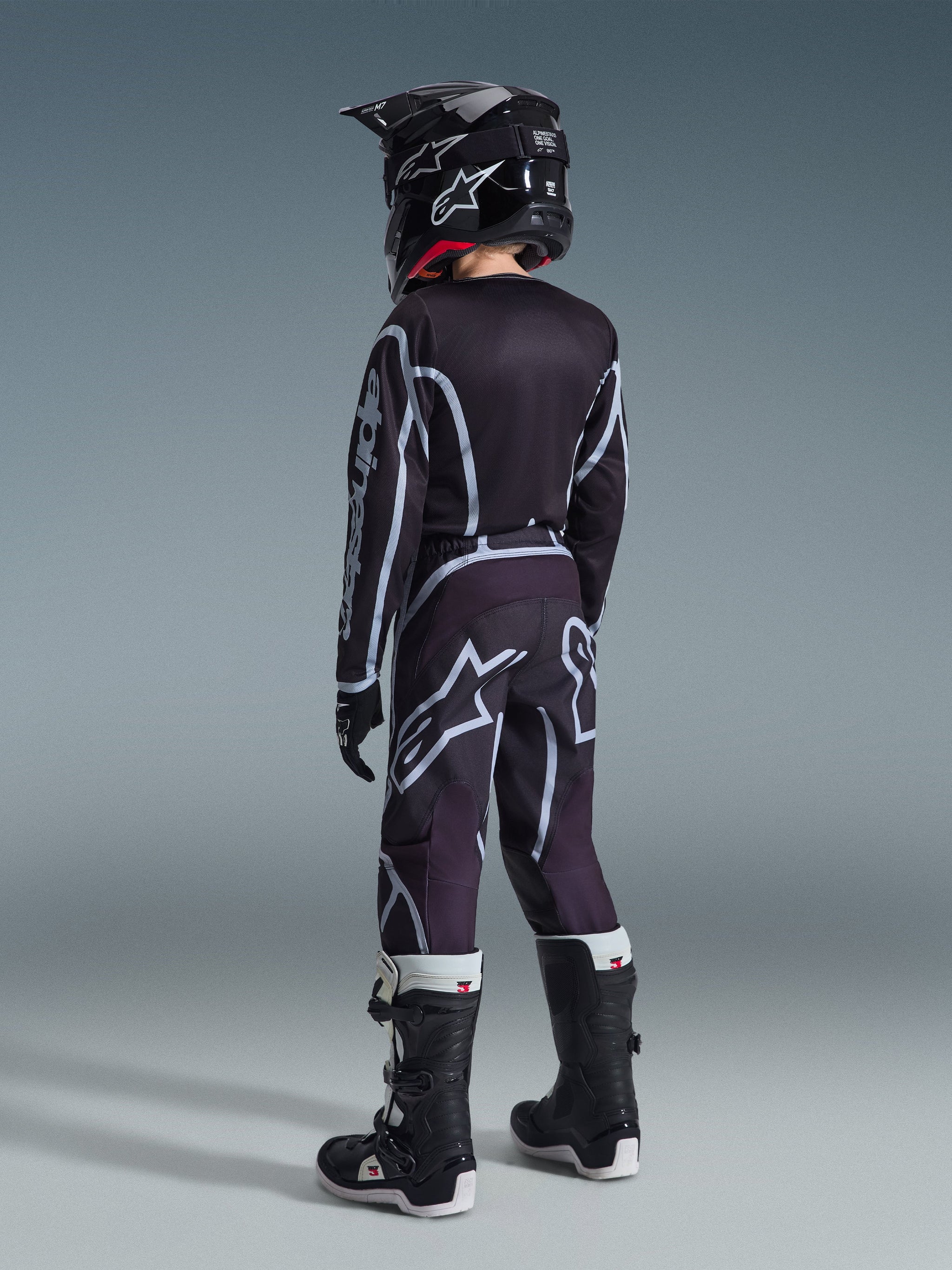 Alpinestars 2026 Youth Fluid Apex Jersey, jeugd motocross shirt, zwart en grijs, gedragen met bijpassende Youth Fluid Grid Pants, zwarte helm en zwart-witte laarzen, achteraanzicht van de volledige off-road race-uitrusting voor jonge rijders