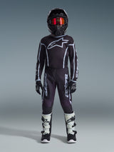 Jonge motorcrossrijder in de Alpinestars 2026 Youth Fluid Apex Jersey in zwart en grijs, gecombineerd met een bijpassende broek, zwarte helm met rood iriserende bril, zwart-witte handschoenen en motorcrosslaarzen, complete uitrustingsset voor jeugd off-road racen