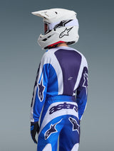 Achteraanzicht van een jonge motorcrosser die de Alpinestars 2026 Youth Fluid Portl Jersey in Ucla Blue White draagt, gecombineerd met een witte Alpinestars helm en bijpassende broek, met grote logo-graphics en een ergonomisch ontwerp voor off-road racen.