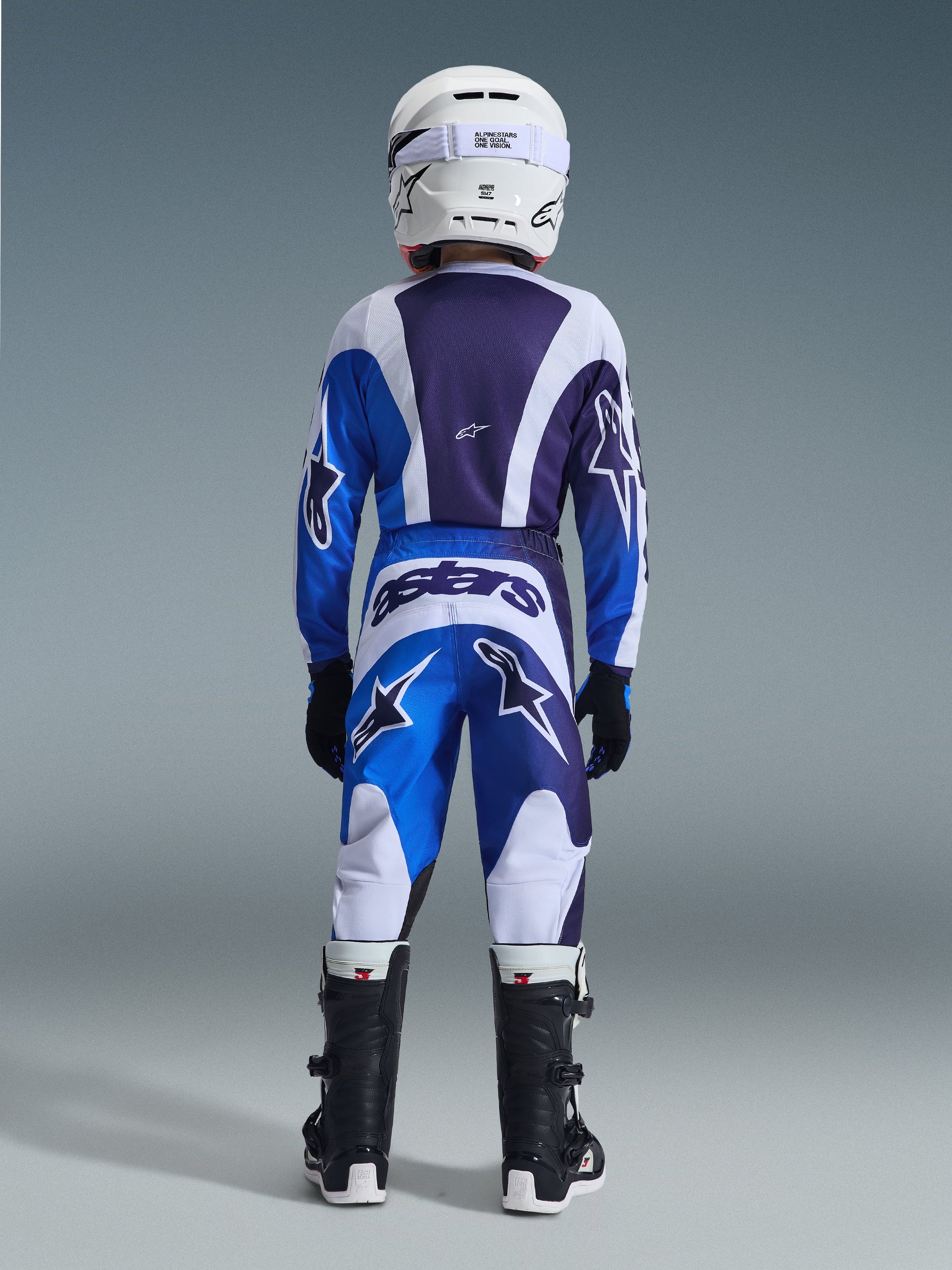 Jeugd motocrossrijder in complete race-uitrusting vanaf de achterkant gezien, Ucla Blue en White Fluid Portl Jersey, bijpassende broek met grote sterlogo's en 'astars' branding, witte helm en zwarte laarzen, gecoördineerde professionele outfit voor off-road rijden.