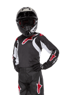 Alpinestars Youth 2024 Racer Lucent Jersey, motorcross jersey, zwart, wit en rood, gedragen door een jeugdrijder met een zwarte helm en bijpassende broek, voorzien van rode sterlogo's en witte grafische accenten, technische off-road kleding voor motorcross