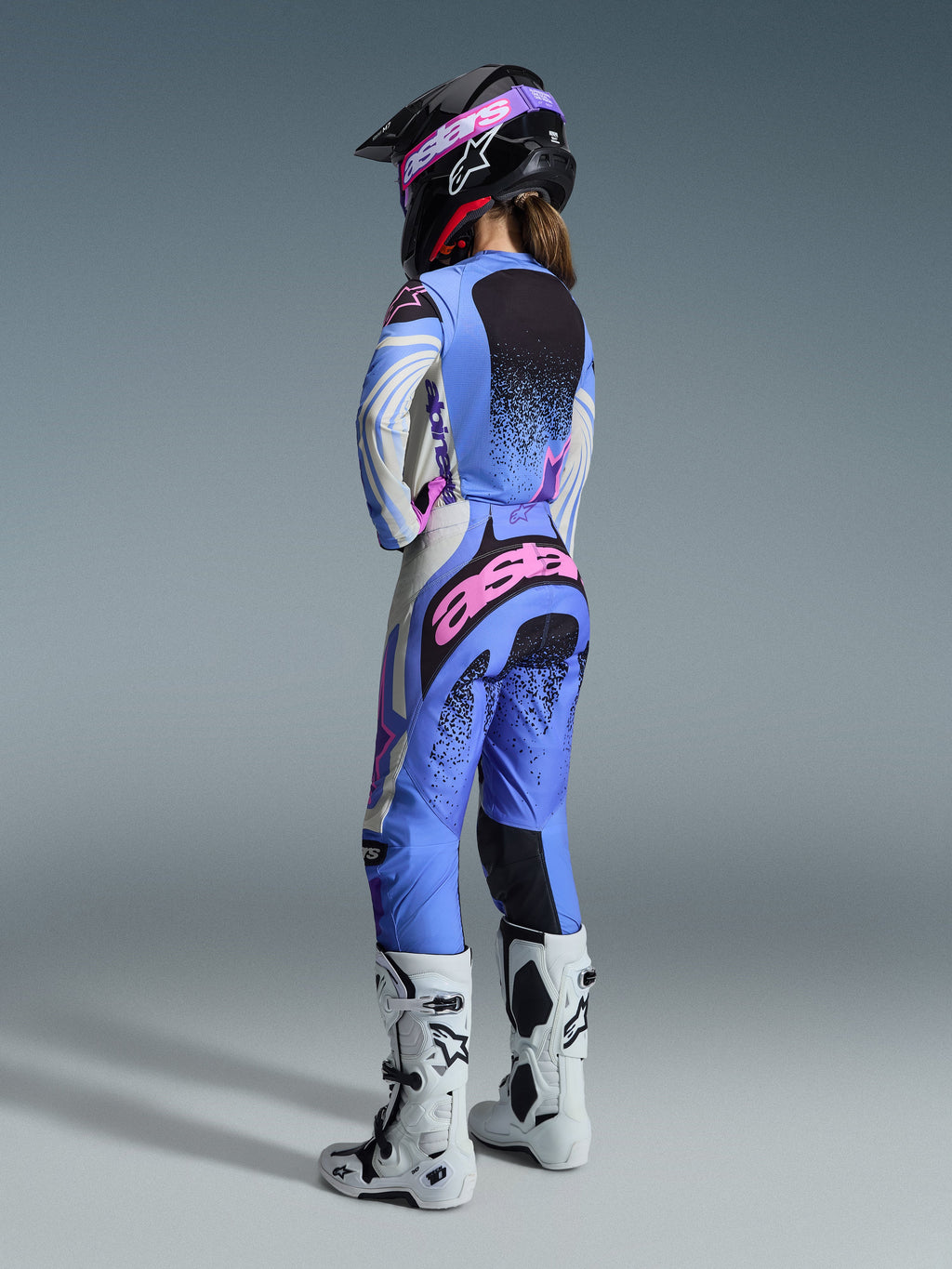 Alpinestars 2026 Stella Techstar Nomur Gear Set, rear view of a female motocross rider wearing a light blue, white, and pink jersey and pants set with black speckled details, paired with a black helmet and white boots, professional racing gear for women.