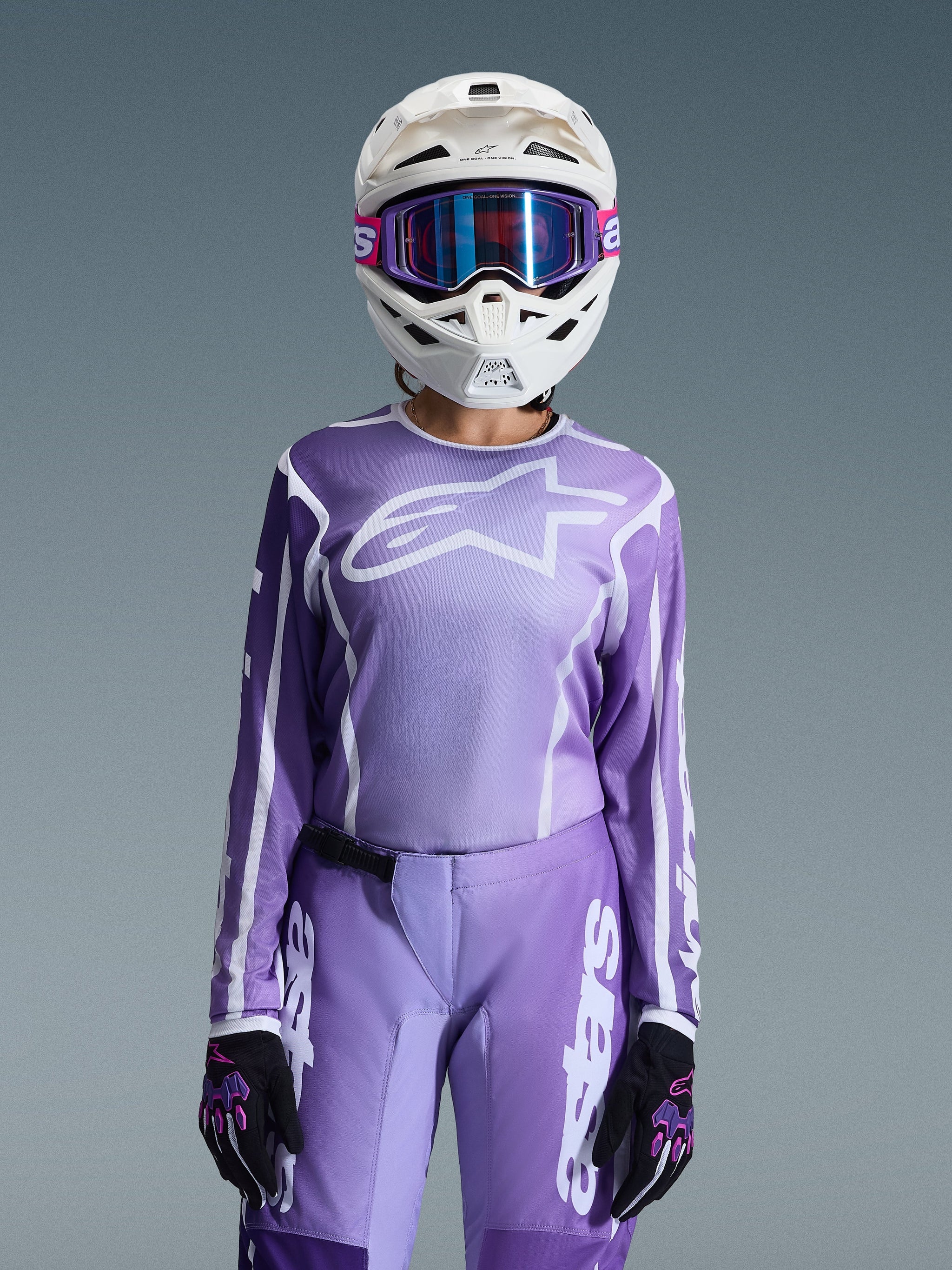 Alpinestars 2026 Woman Stella Fluid Apex Jersey in Violet White, worn by a female rider with matching purple Stella Fluid Apex pants, white helmet with purple goggles, and black/purple gloves, coordinated off-road racing gear