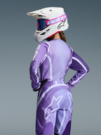 Motocross rider wearing Alpinestars 2026 Stella Fluid Apex Gear Set, Purple and lavender jersey and pants with white graphics, white helmet with pink and purple goggles, dedicated women's fit, coordinated gear for off-road racing