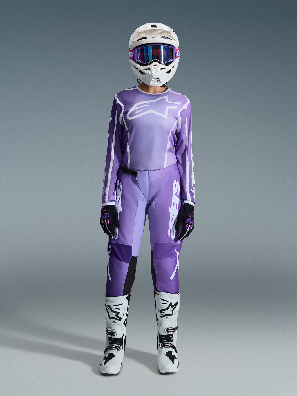 Motocross rider wearing the Alpinestars 2026 Stella Fluid Apex Gear Set, purple jersey with white logo, matching purple pants with white branding, white helmet with pink goggles, white and black boots, coordinated women's motocross outfit for off-road racing