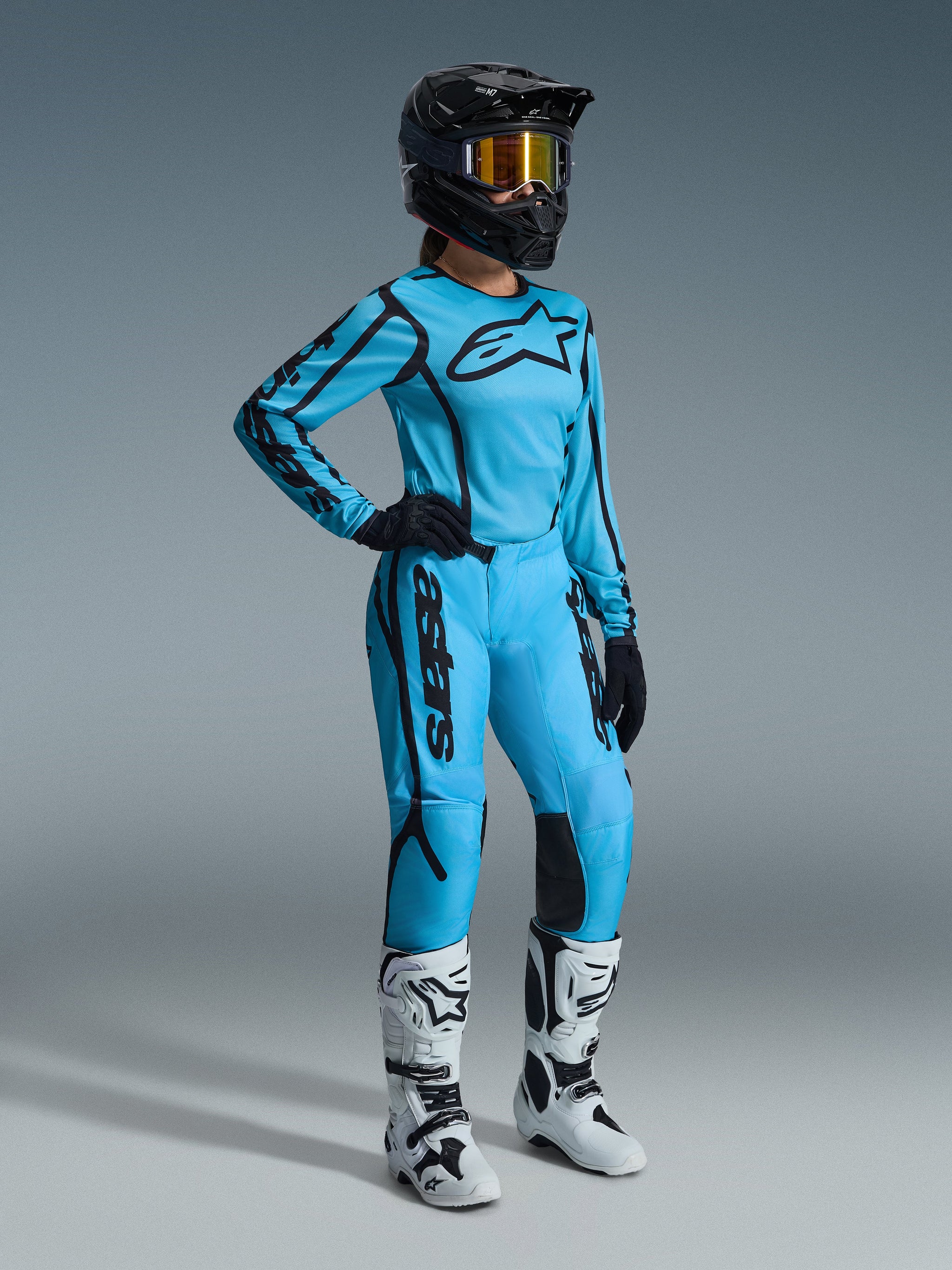 Alpinestars 2026 Woman Stella Fluid Apex Jersey, light blue and black motocross jersey, worn as a complete racing gear set with matching pants, black helmet with gold-tinted goggles, and white boots, optimized for women's off-road performance.