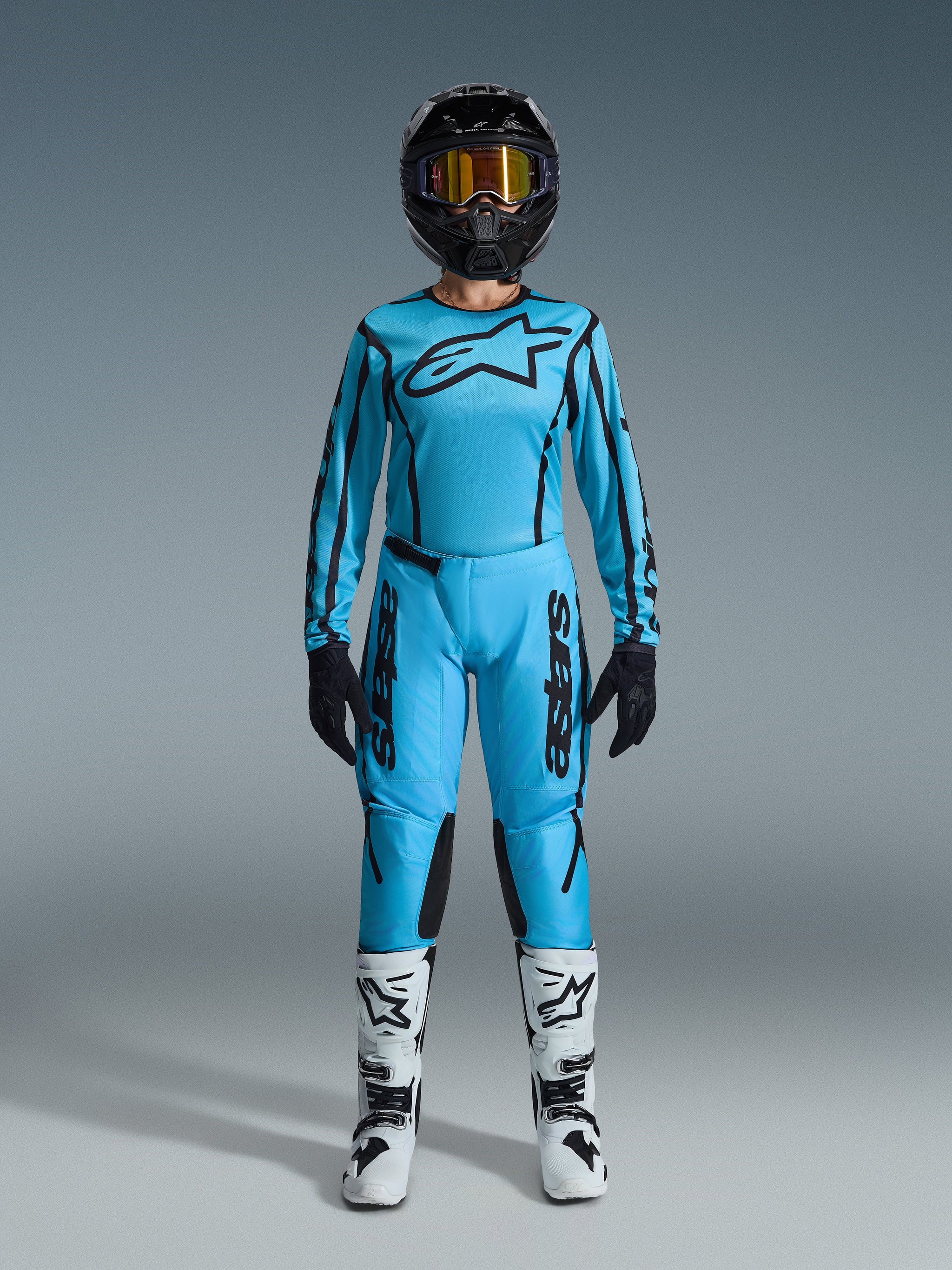 Motocross rider wearing the 2026 Alpinestars Stella Fluid Apex Jersey and matching pants in light blue with black accents, black helmet with gold goggles, white and black boots, and black gloves, complete women's off-road gear set.
