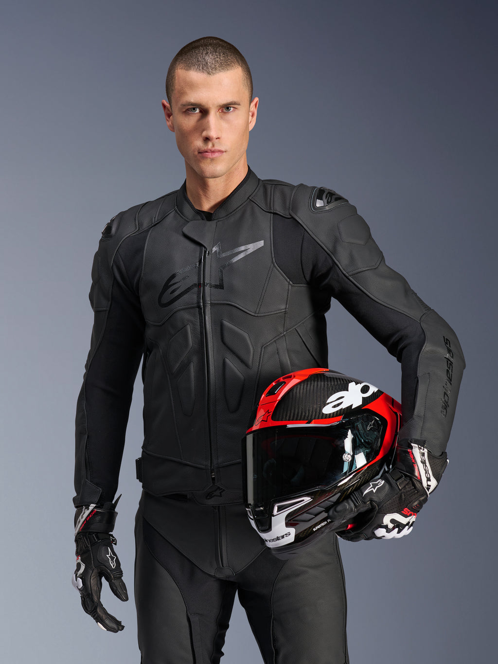 Alpinestars Dusk Leren Jas, zwarte motorjas, premium leren constructie met gewatteerde bescherming, mannelijk model houdt rood-zwarte racehelm vast, draagt bijpassende leren broek en handschoenen, professionele motorsportuitrusting
