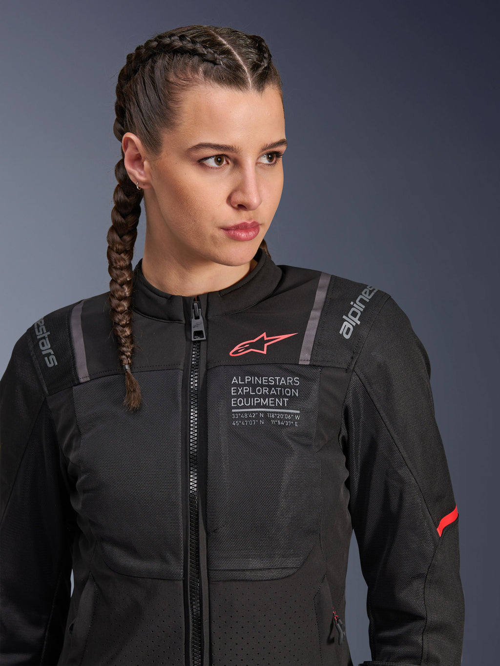 Alpinestars Stella ST-2 Air Jacket, dames motorjas in zwart, voorzien van mesh-panelen voor luchtstroom, 'Alpinestars Exploration Equipment' branding met coördinaten, rood logo-accent op de borst, model met gevlochten haar